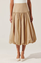 A beige high-waisted skirt with a gathered, voluminous, balloon-style silhouette that falls to mid-calf length. The skirt features a fitted waistband and gathered fabric creating pleats and a bubbly effect at the hem. The wearer pairs it with white pointed-toe heels.